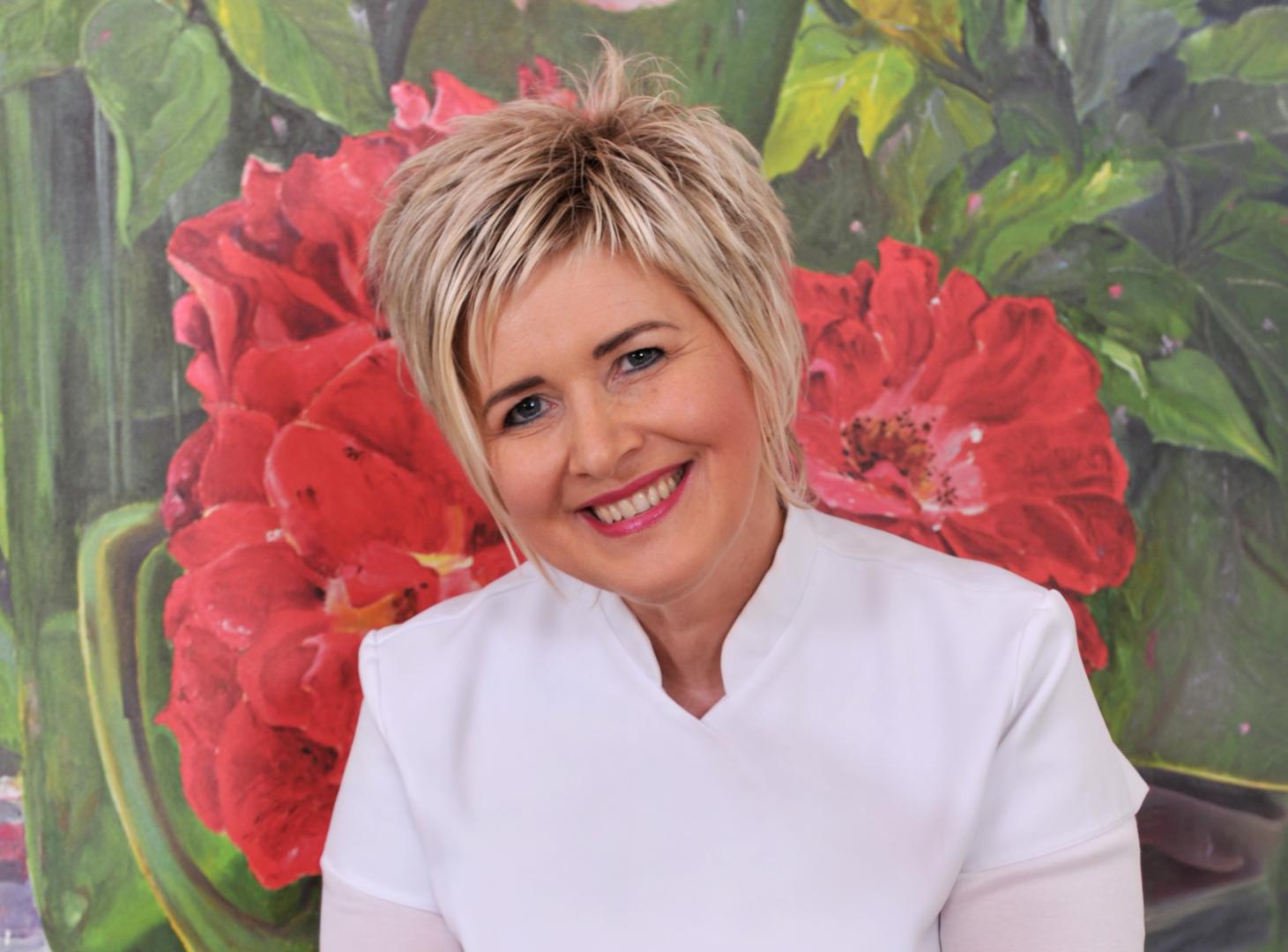 Sylvia Scherrer mit kurzem blondem Haar und weißer Bluse vor einem Gemälde mit großen roten Blumen